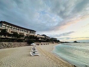 Una playa con tumbonas alineadas, un edificio de varios pisos con balcones y un mar tranquilo.