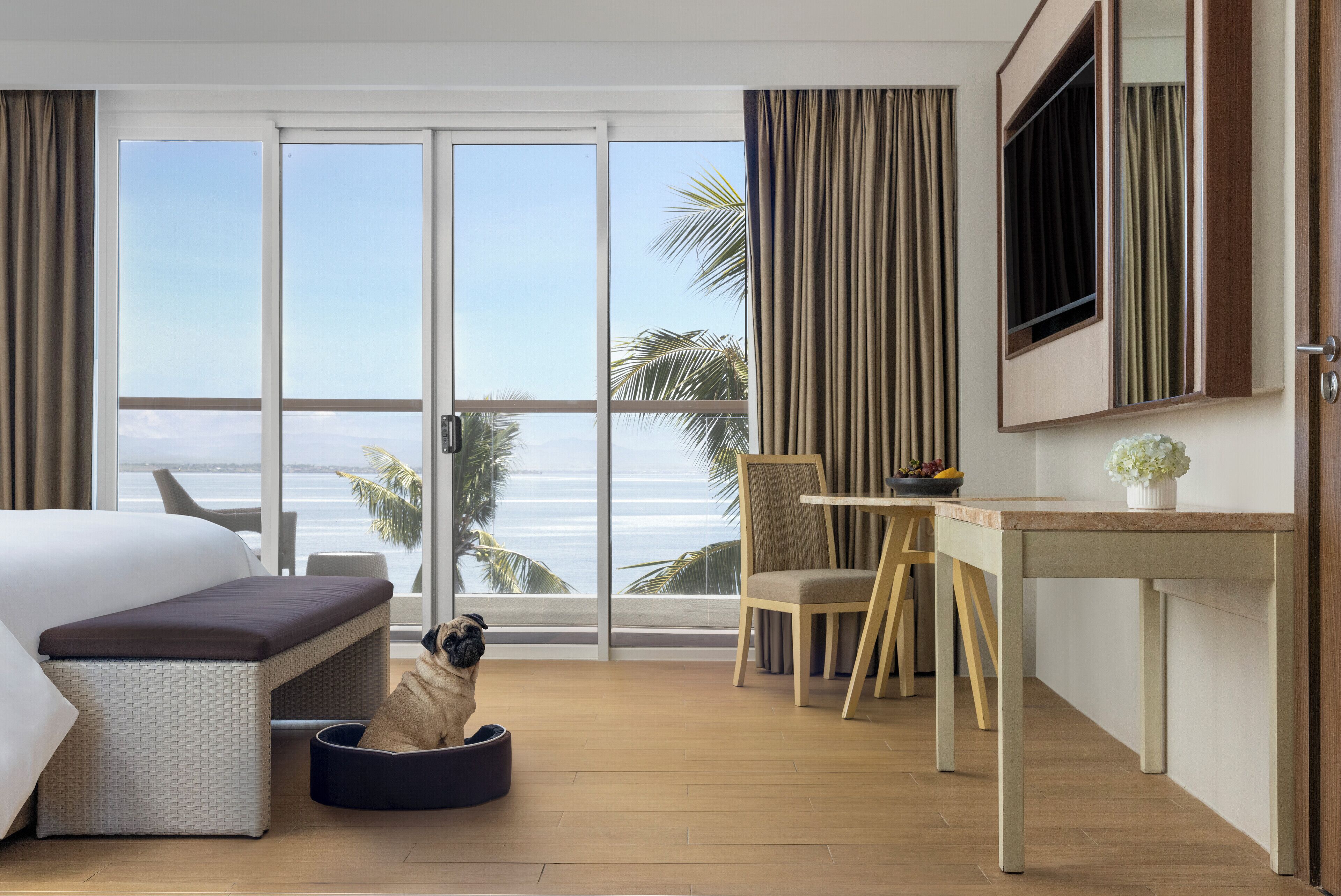 Quarto de hotel com cama, escrivaninha, cadeira, televisão e uma grande janela com vista para o mar.