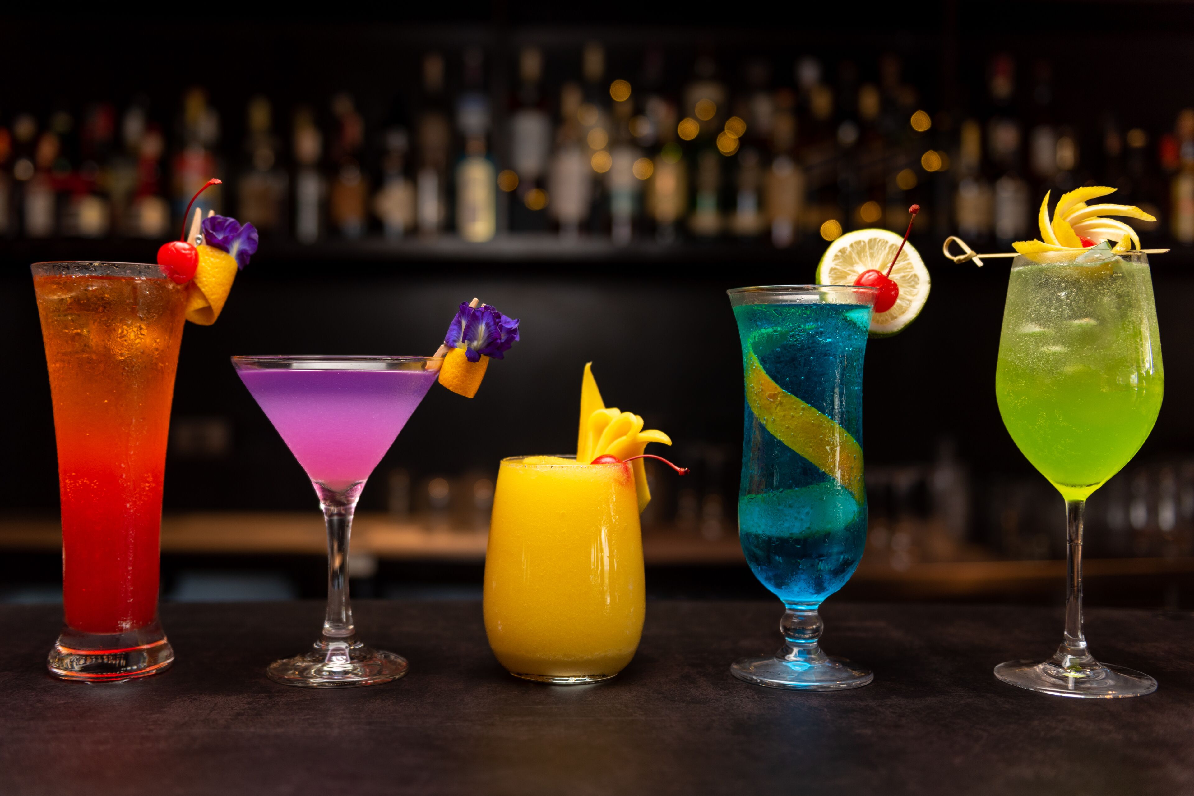 Un bar con cinco cócteles coloridos, cada uno con una guarnición original.