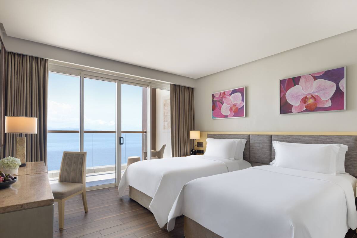 Ein Hotelzimmer mit zwei Betten, einem Schreibtisch mit Stuhl, Meerblick und zwei Orchideenbildern an der Wand.
