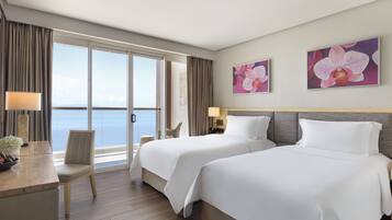 A hotel room with two beds, a desk with a chair, a view of the sea, and two orchid paintings on the wall.