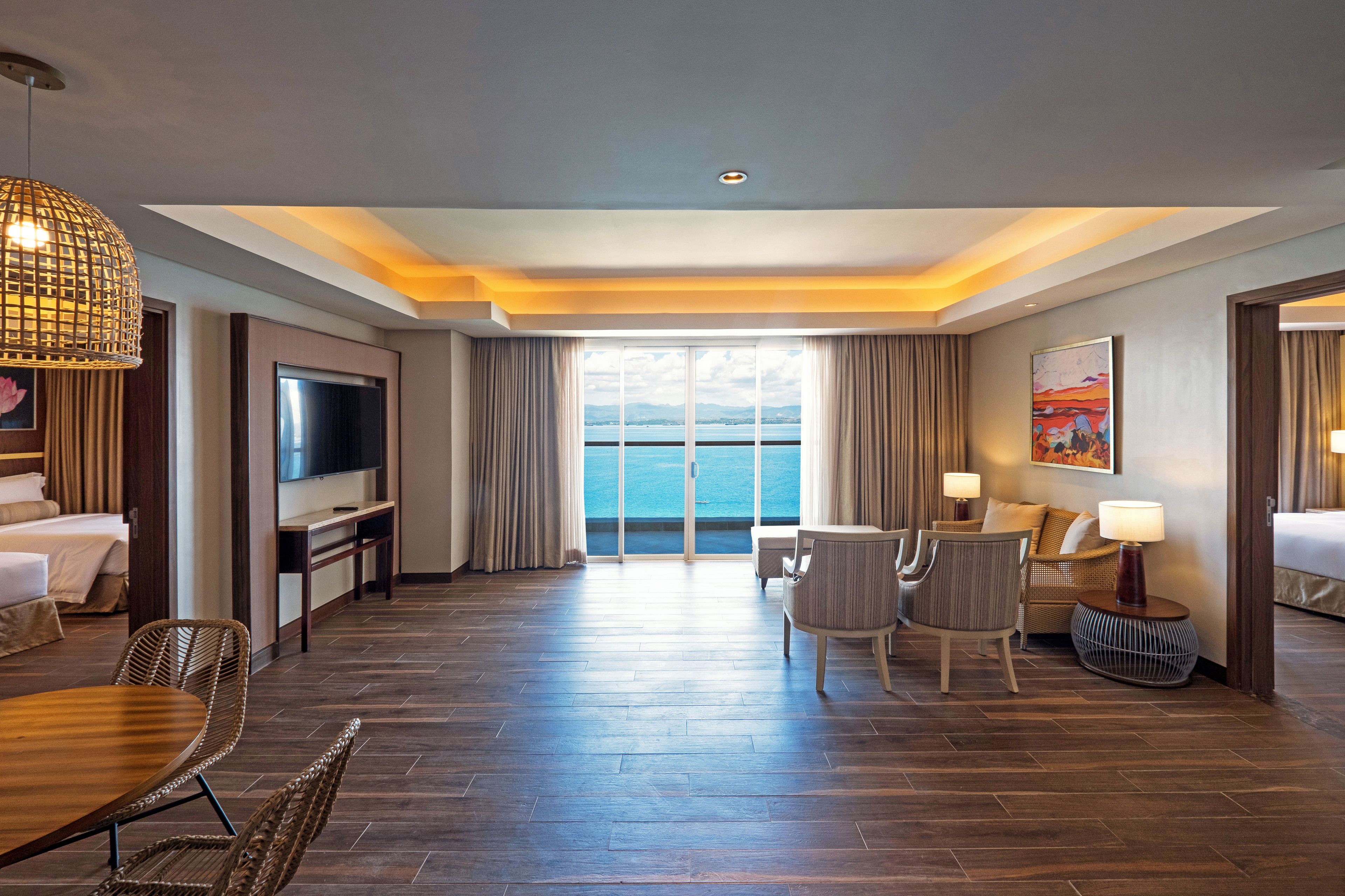 Une chambre d’hôtel dotée d’une grande télévision, d’une table à manger, de deux chaises, d’un canapé, d’une lampe et offrant une vue sur la mer.
