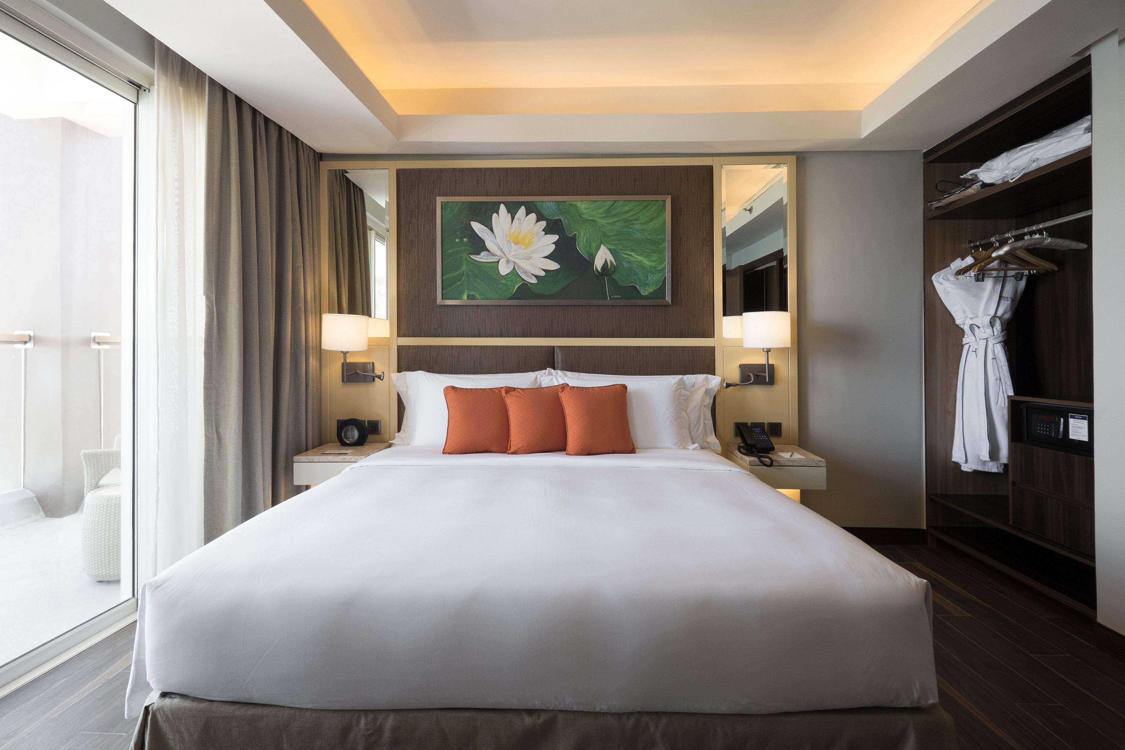 A hotel room with a large bed, a painting of a flower, a wardrobe, and a bathroom visible through a glass door.
