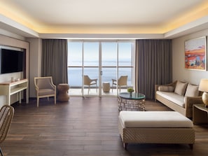 A modern hotel room with a large sliding glass door to a balcony, a flat-screen TV, a sofa, a coffee table, and wicker furniture.