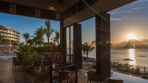 Un vestíbulo de hotel con vistas a la puesta de sol sobre el agua, mobiliario exterior con sillas y mesas, y un edificio grande al fondo.