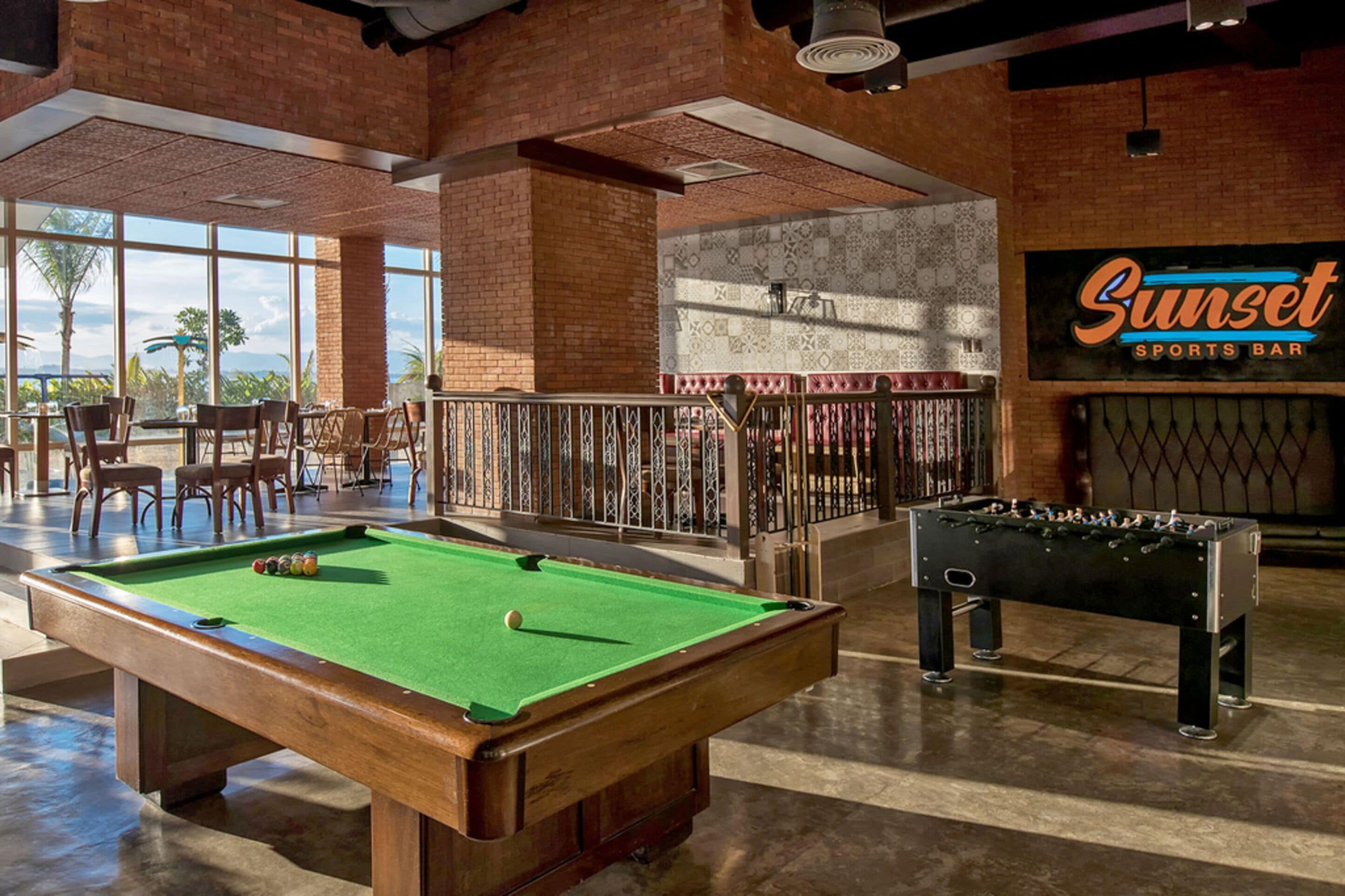 A pool table and foosball table in a sports bar with a view of palm trees.