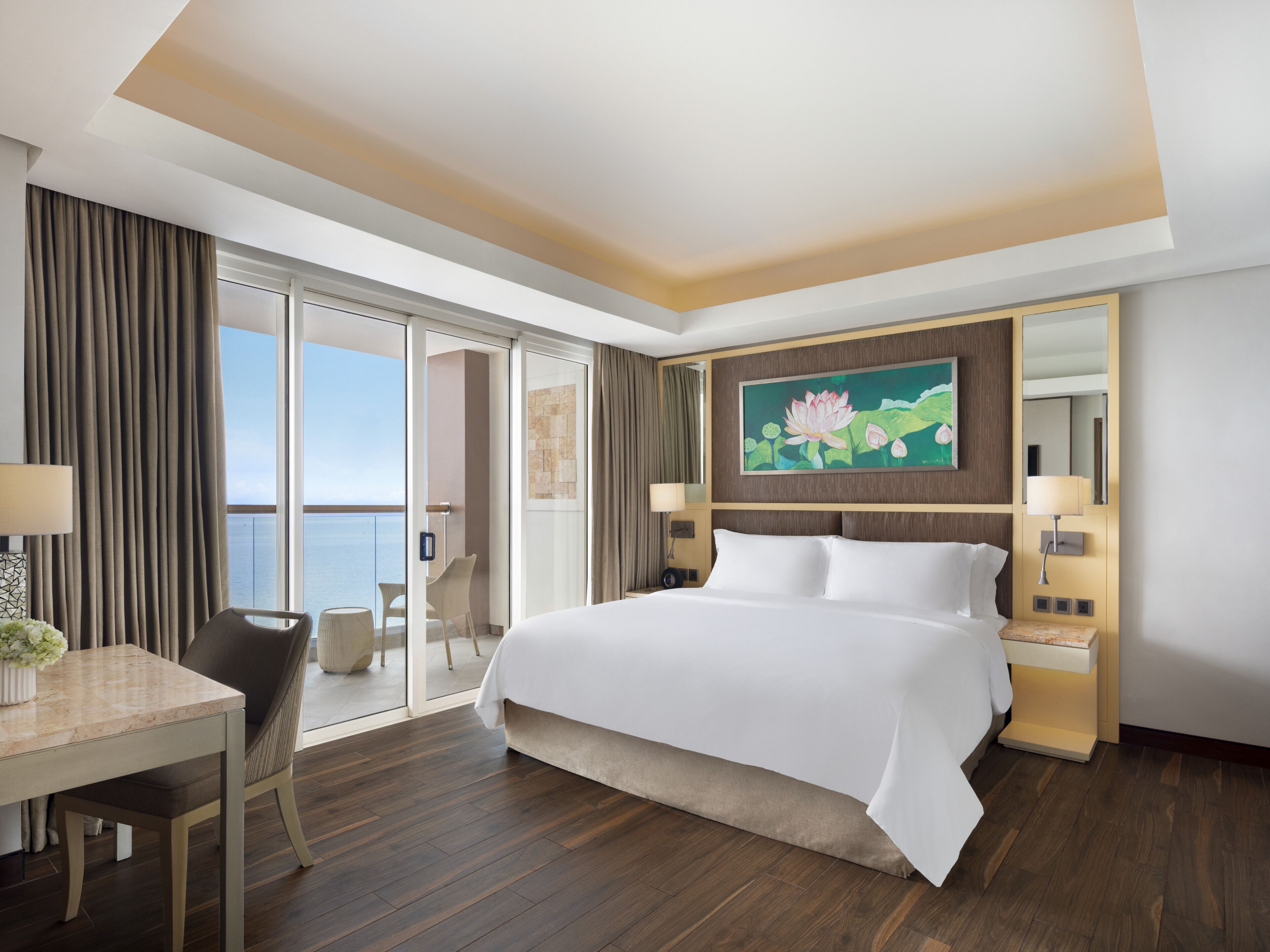 A hotel room with a large bed, a desk, a chair, a balcony with a view of the sea, and a painting on the wall.