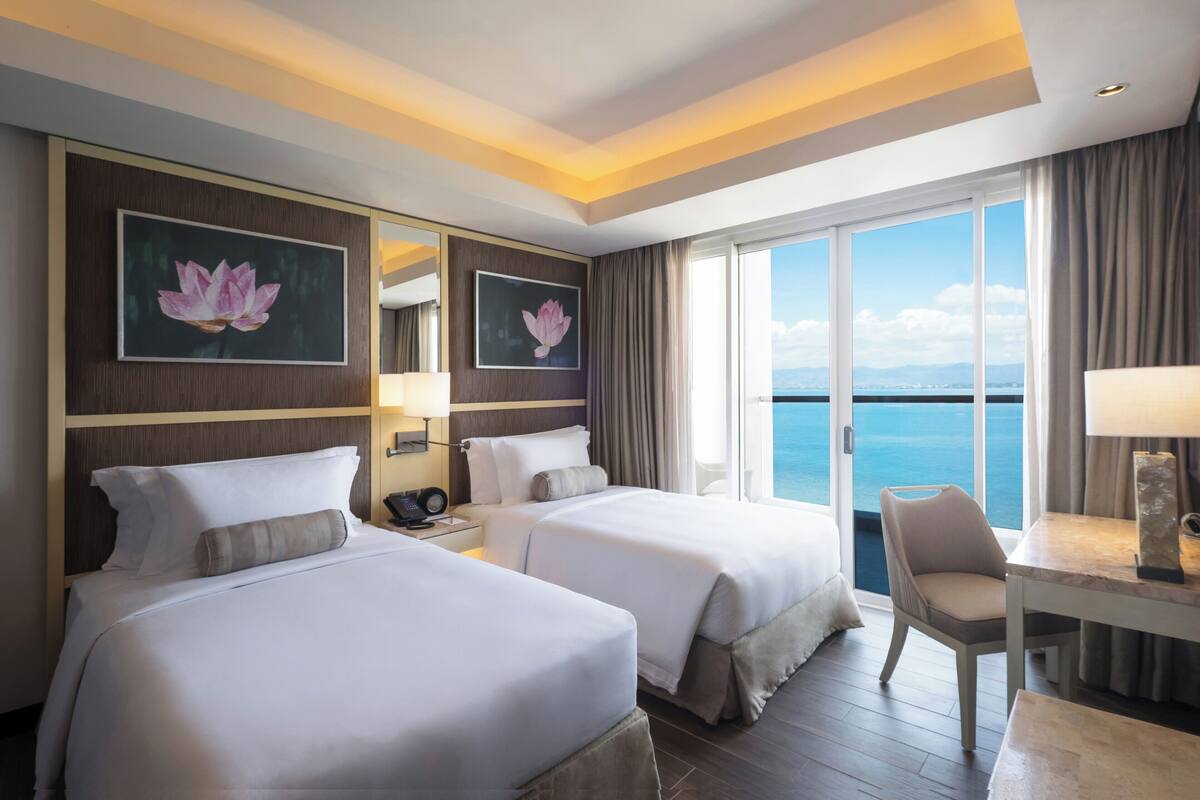 Ein Hotelzimmer mit zwei Betten, einem Schreibtisch, einem Stuhl, einem großen Fenster mit Meerblick und Bildern an den Wänden.