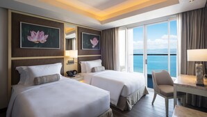 Habitación de hotel con dos camas, un escritorio, una silla, un ventanal con vistas al mar y obras de arte en la pared.