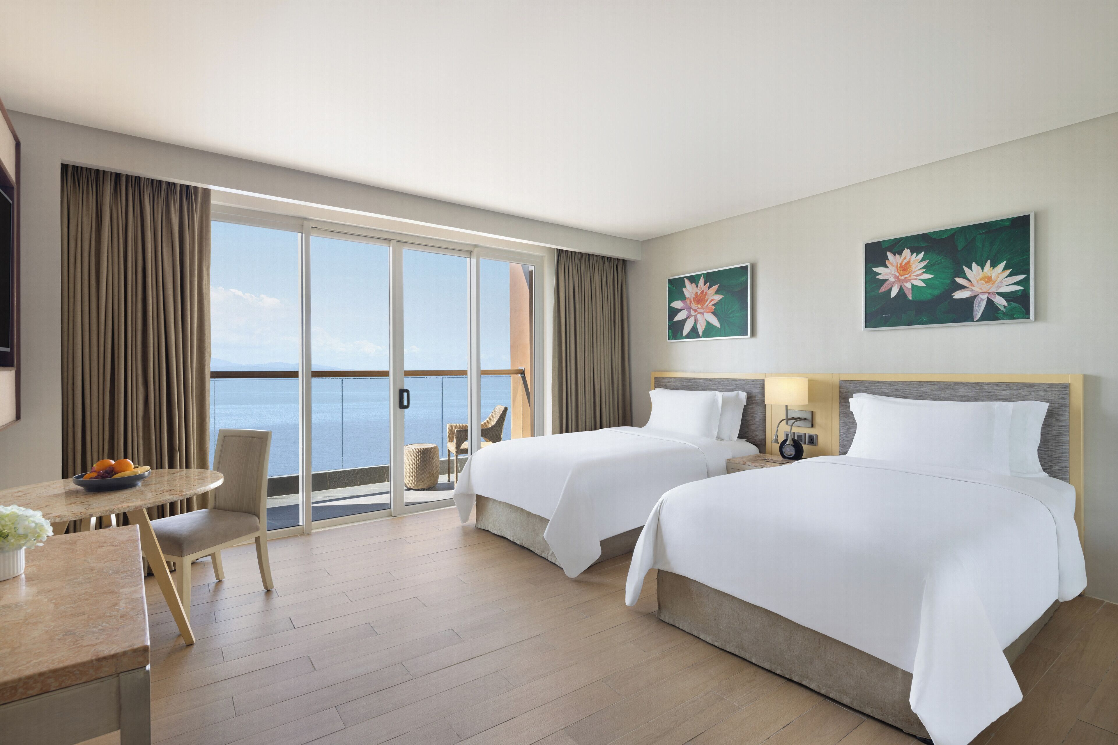 Habitación de hotel con dos camas, una mesa de comedor con fruta, una silla, vista al mar y dos cuadros en la pared.