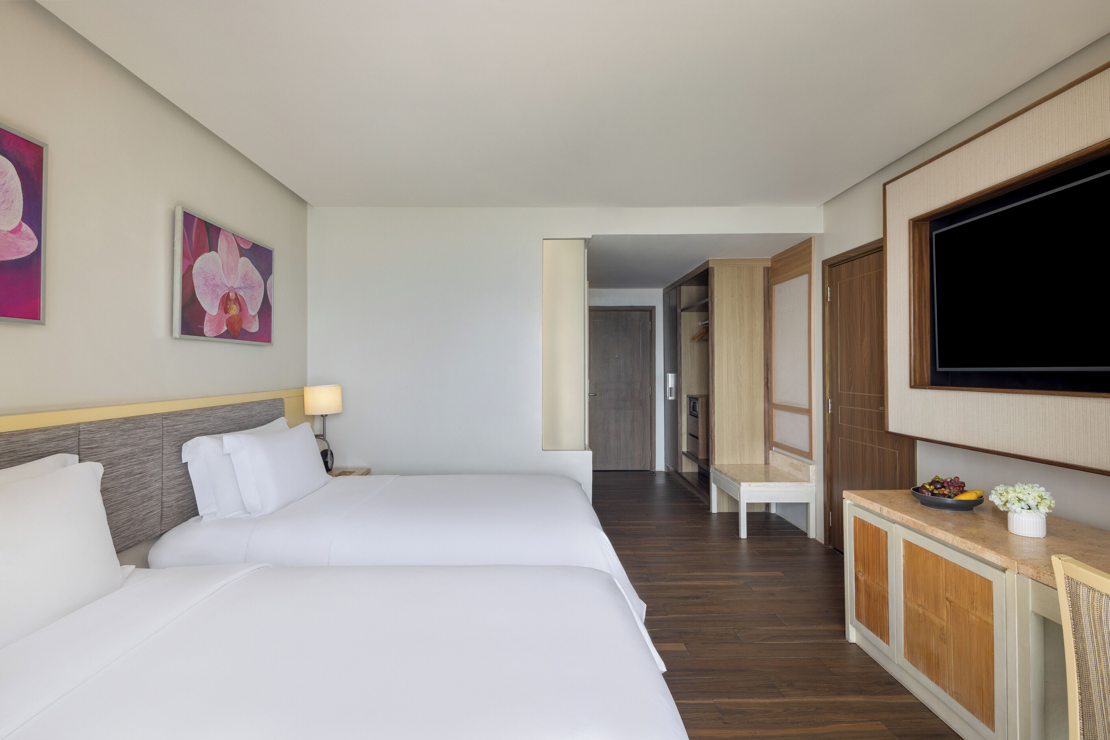 A hotel room with two beds, a TV, a desk with a bowl of fruit, and a door leading to another room.