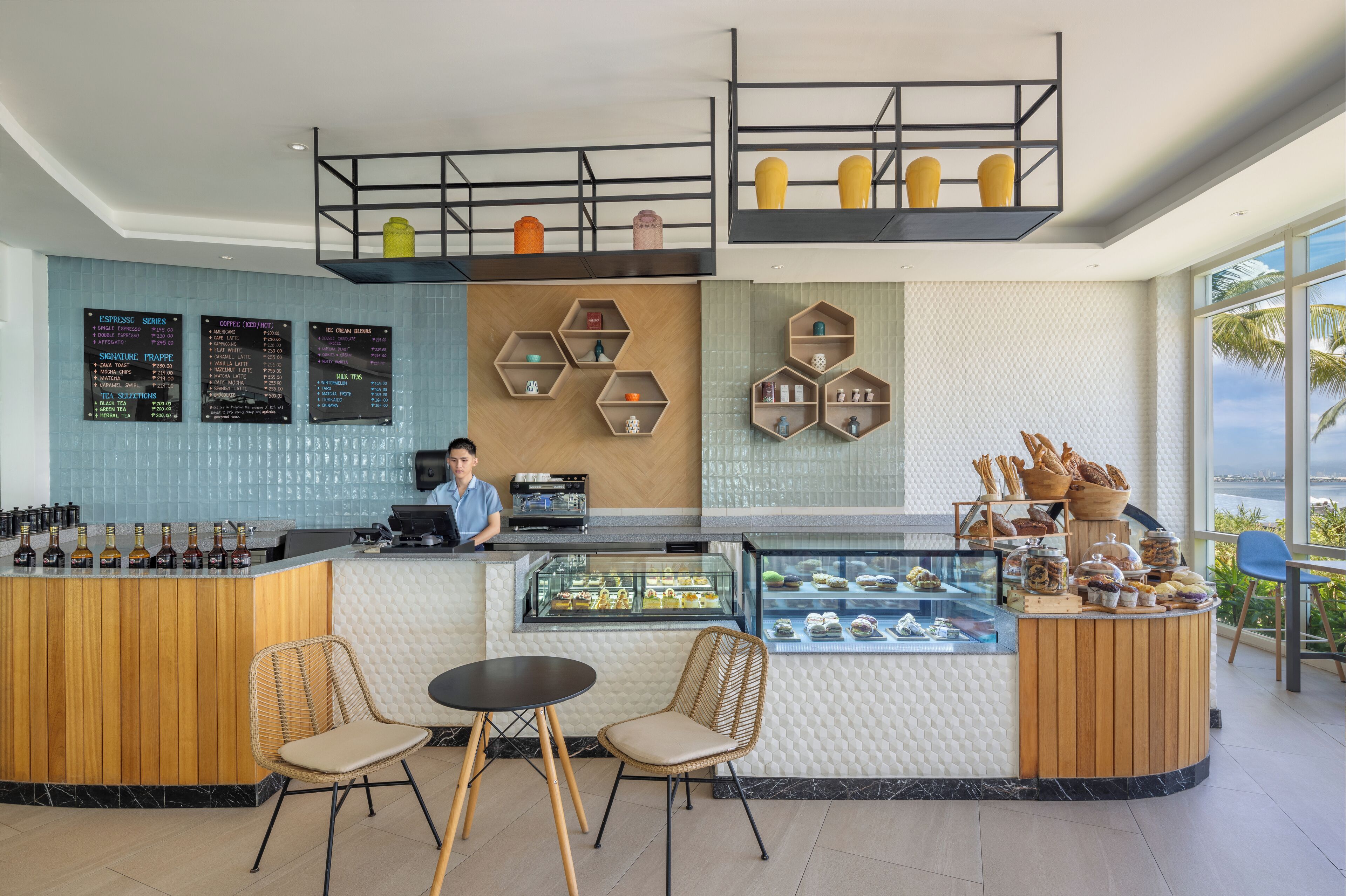 A modern cafe with a wooden counter, menu boards, hexagonal shelves, and a view of the ocean.