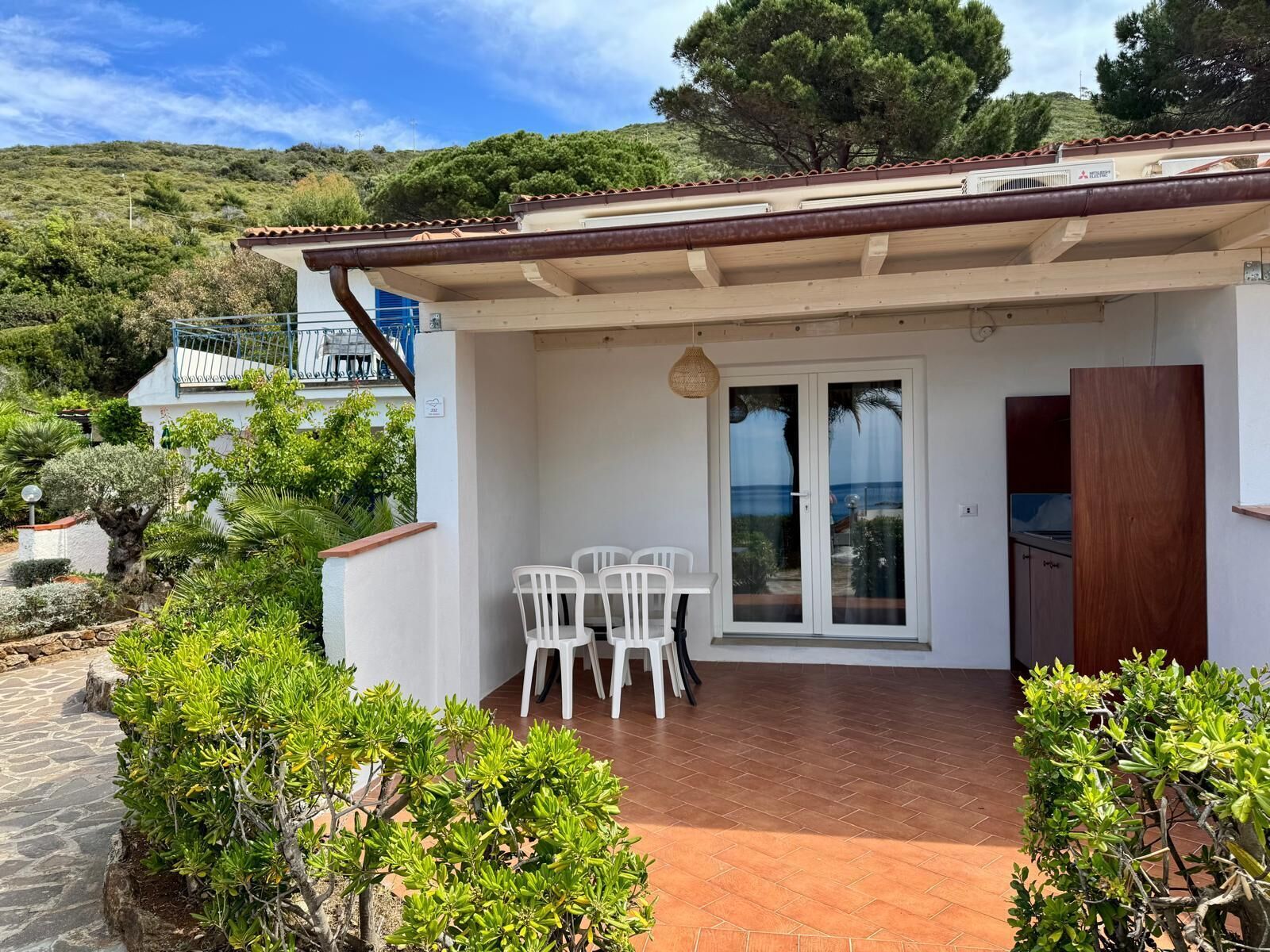 A small, white building with a tiled roof, a patio with a table and chairs, and a view of a hillside.
