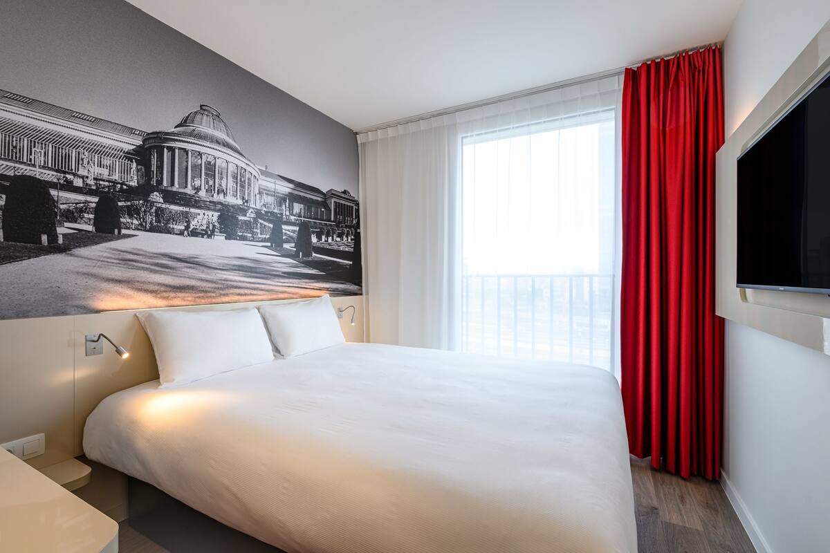 Ein Hotelzimmer mit einem großen Bett, einem an der Wand montierten Fernseher, einem Fenster mit roten Vorhängen und einem Wandbild eines Gebäudes.