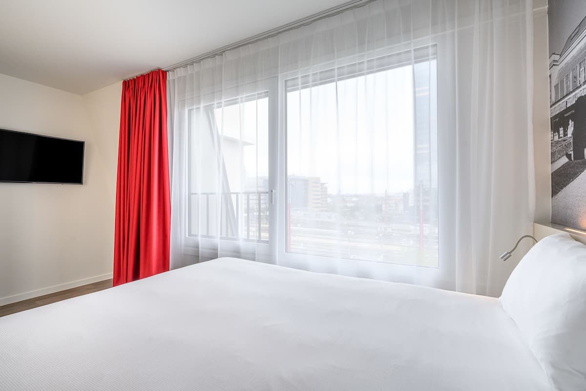 Ein Hotelzimmer mit einem großen Bett, einem flachbildfernseher an der Wand und einem Fenster mit durchsichtigen Vorhängen.