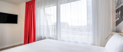 A hotel room with a large bed, a flat-screen TV mounted on the wall, and a window with sheer curtains.