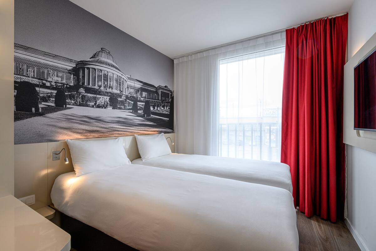 Ein Hotelzimmer mit zwei Betten, einem großen Fenster mit roten Vorhängen und einem Wandgemälde eines Gebäudes.