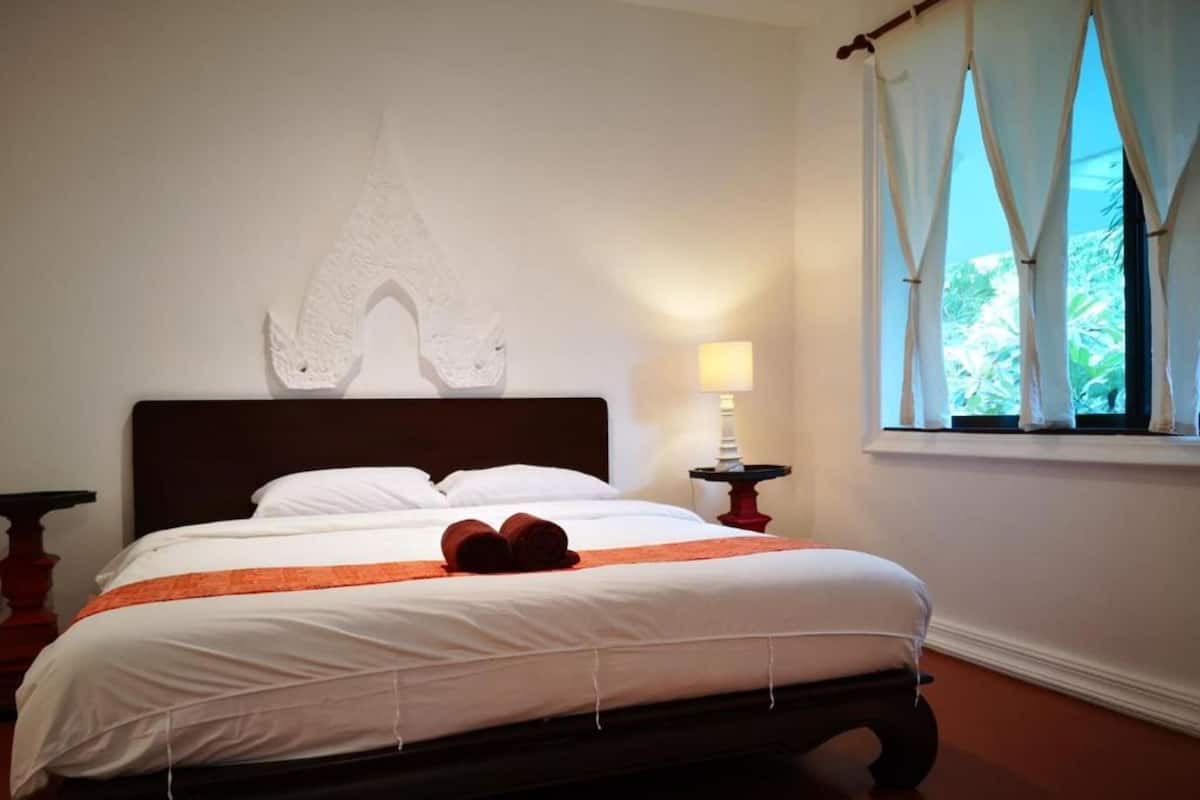 A neatly made bed with white linens and an orange accent, a wall-mounted decorative piece, a bedside table with a lamp, and a window with curtains.