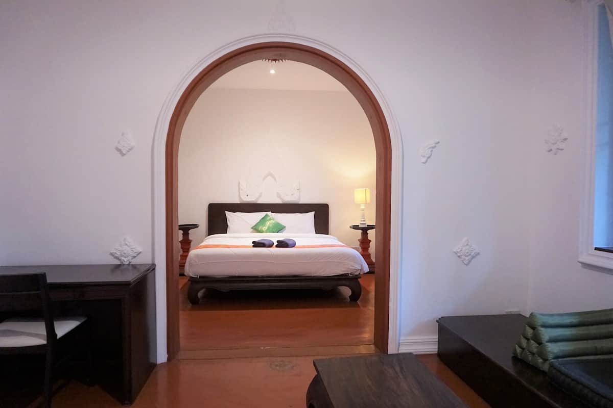 A hotel room with a wooden floor, a bed with white bedding and a green pillow, a small table with a lamp, and a sofa.