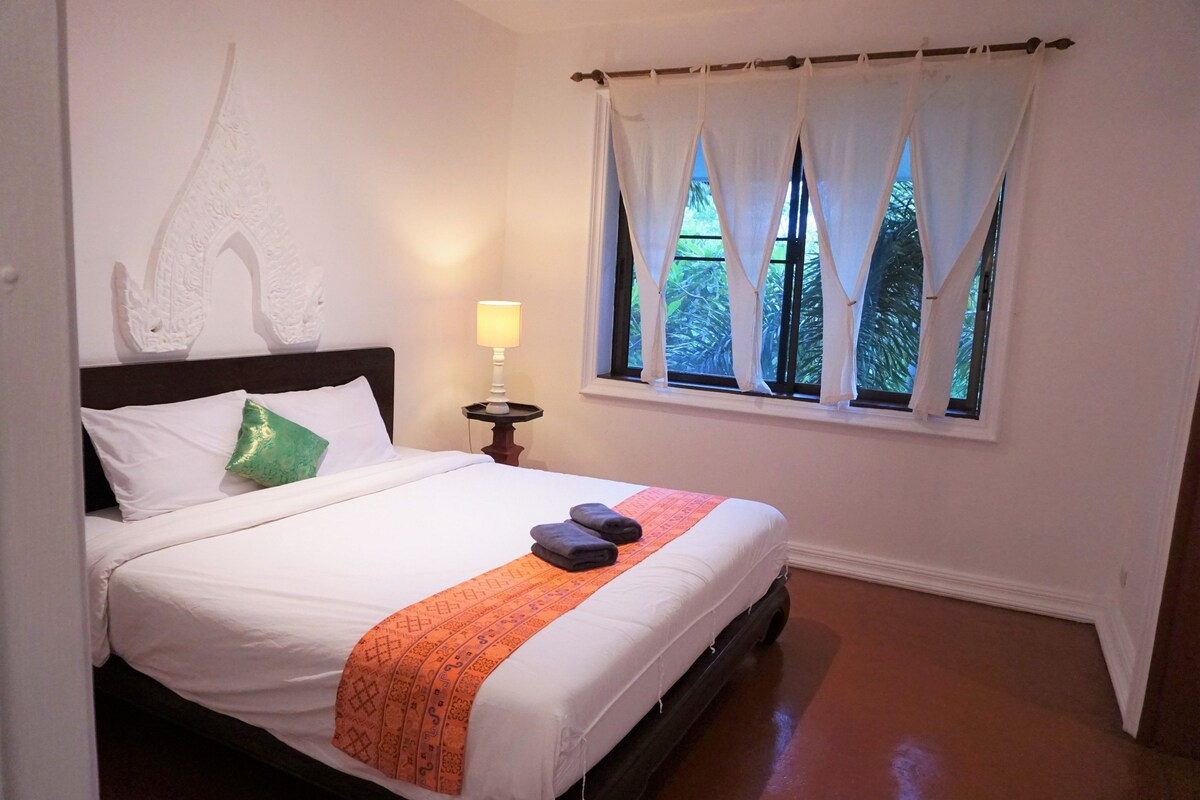 A neatly made bed with white linens, a green pillow, and an orange blanket. There is a bedside lamp, a window with white curtains, and a wall-mounted artwork.