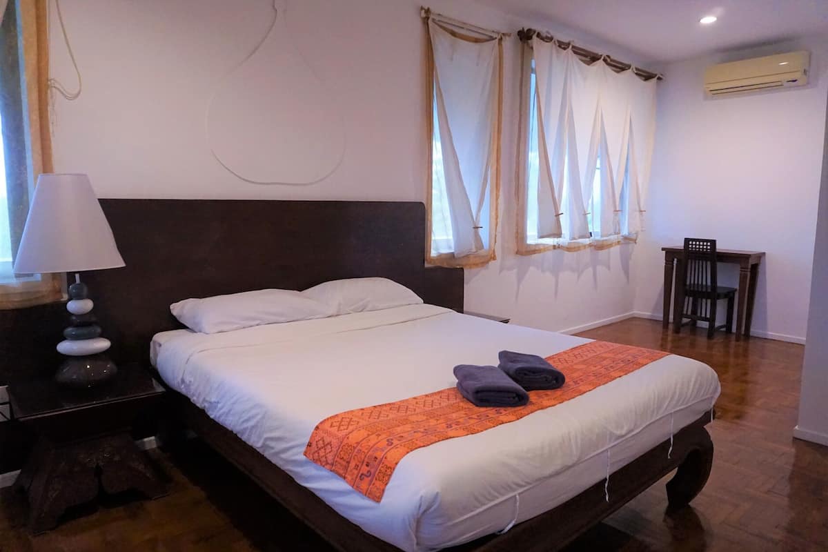 A neatly made bed with a white comforter and an orange decorative throw. There is a wooden nightstand with a lamp and a small table beside the bed.