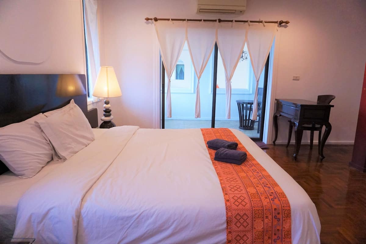 A neatly made bed with white linens and an orange patterned throw, a bedside lamp, and a view of the outside balcony through sheer curtains.