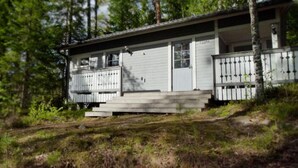Eine kleine, eingeschossige Holzhütte mit Veranda und umzäunter Garten, umgeben von Bäumen.