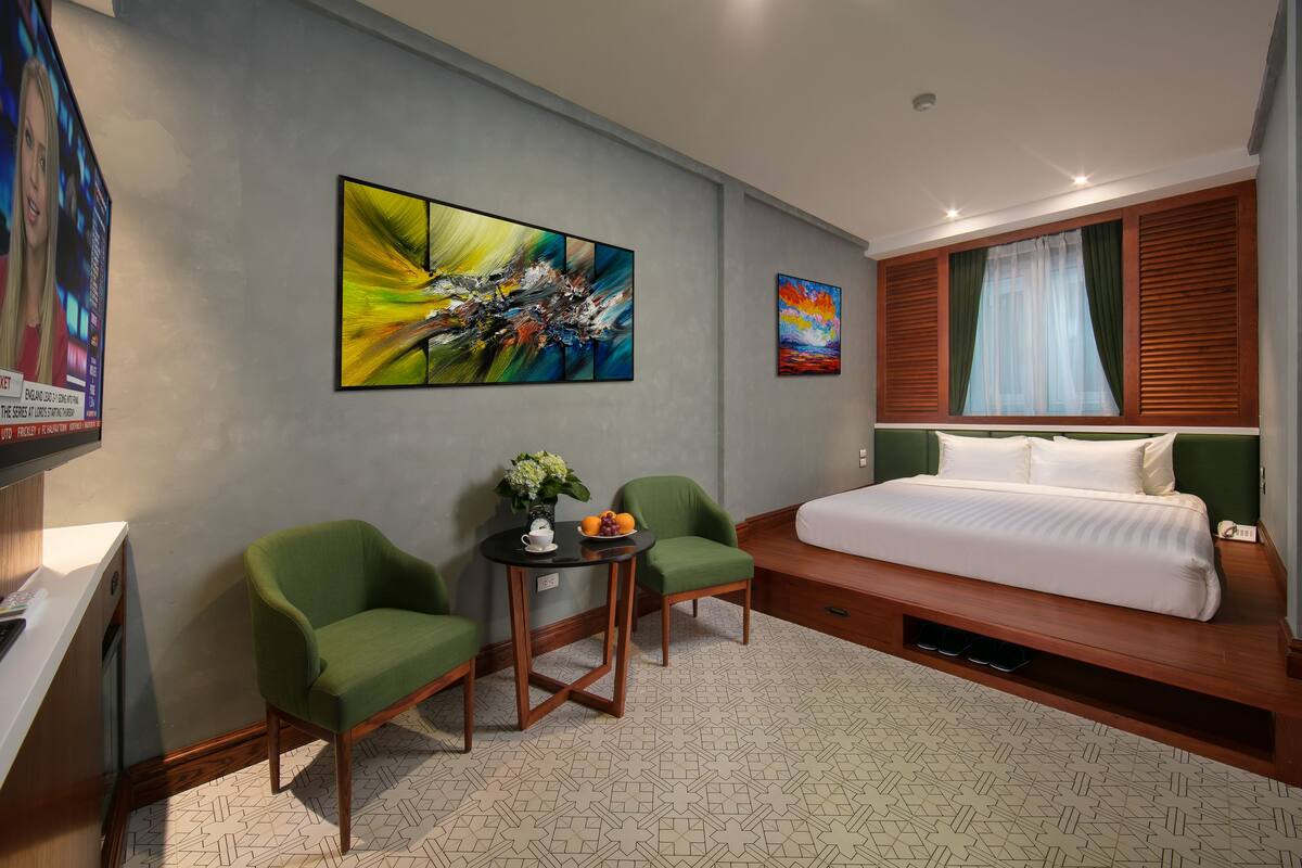 A modern hotel room with a large bed, two green armchairs, a small table with a vase and fruit, and abstract paintings on the walls.