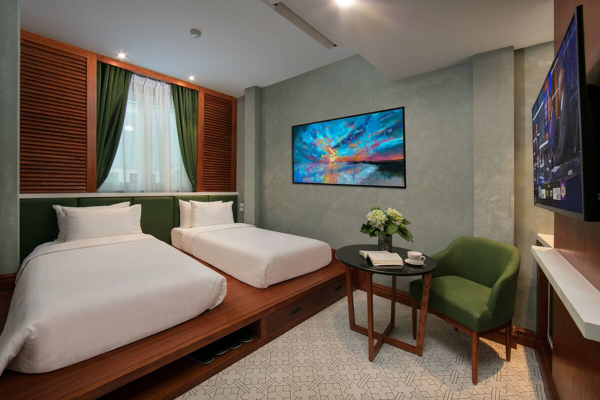A hotel room with two beds, a TV, a painting, and a small table with a chair.