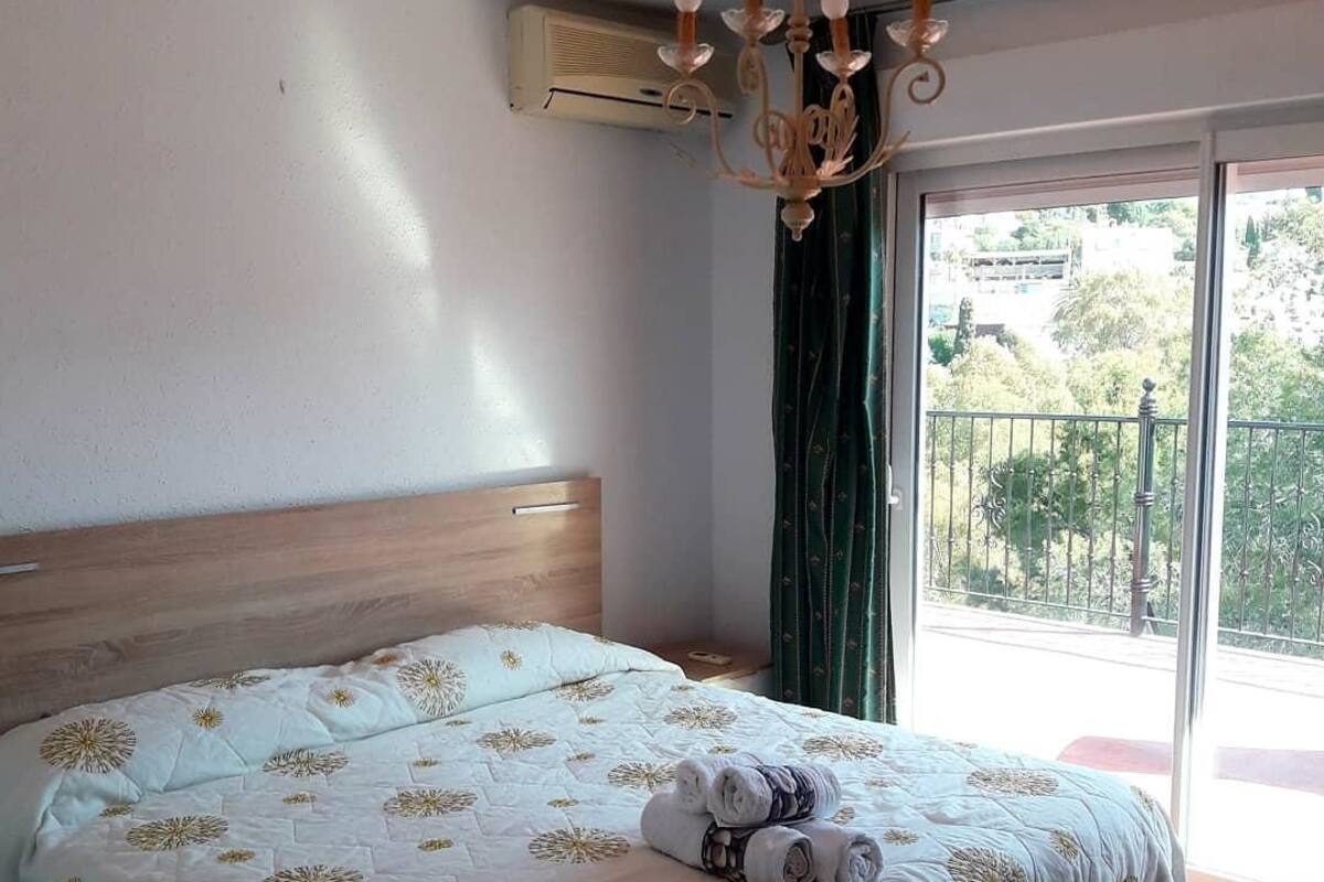 A hotel room with a large bed, a wooden headboard, a chandelier, an air conditioning unit, and a balcony with a view.