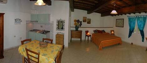 Una habitación con un armario de madera, una mesa de comedor con sillas, una kitchenette y una cama con colcha a rayas, además de una ventana con cortinas azules.