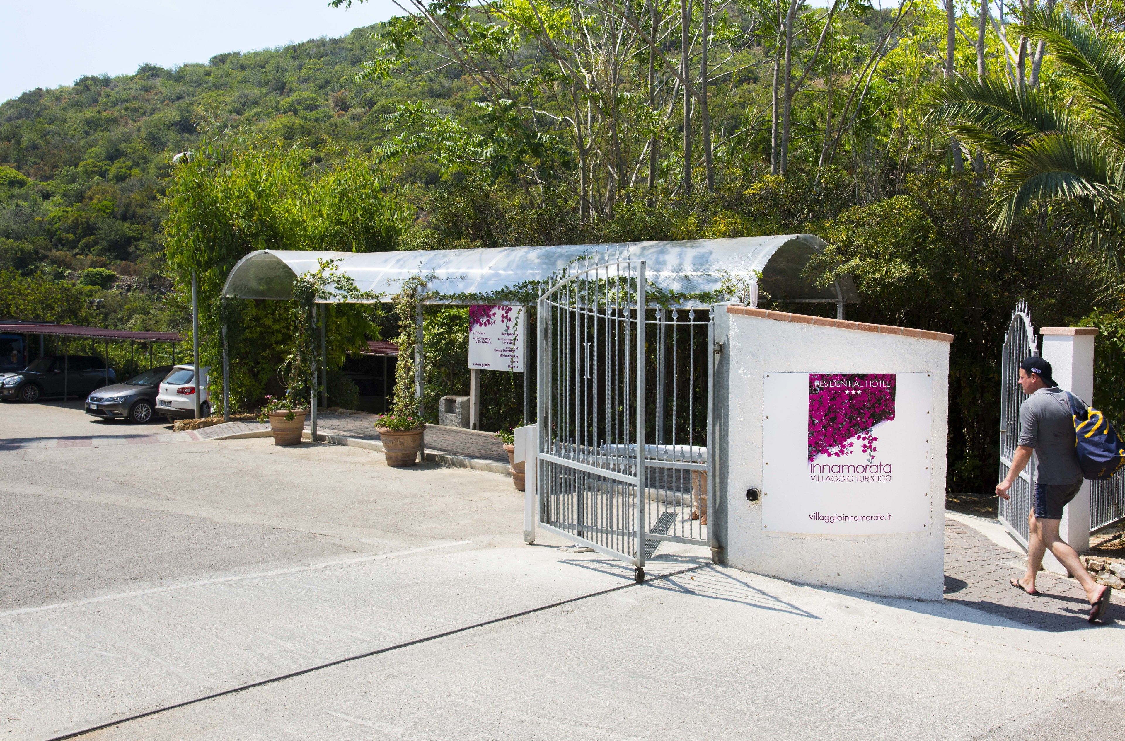 Une entrée fermée par un portail mène à un hôtel résidentiel doté d’un espace d’attente abrité, entouré d’une végétation luxuriante et bordé par des collines.