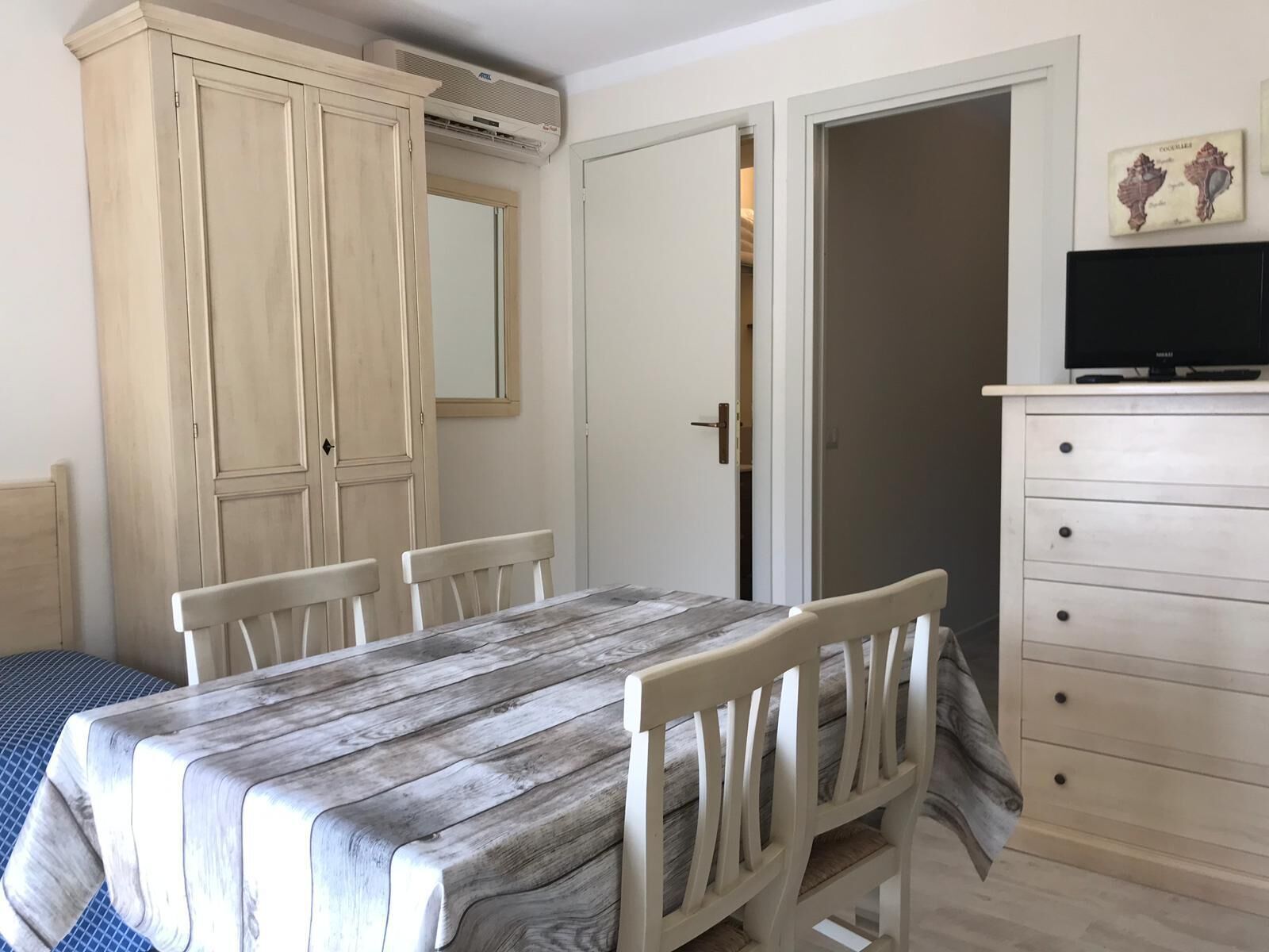 Une pièce avec une table à manger en bois, des chaises blanches, une commode blanche et un téléviseur posé sur le dessus.