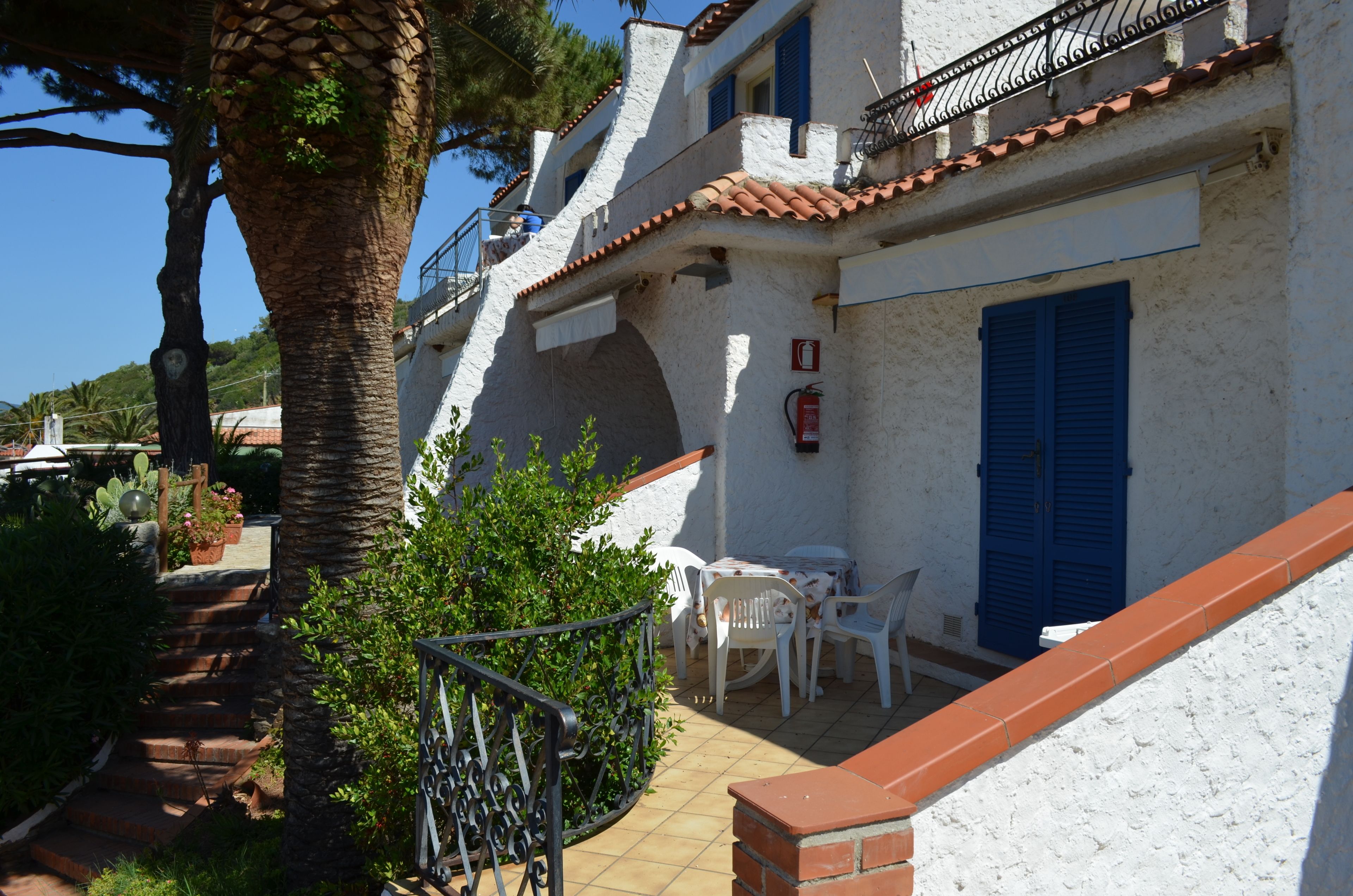 A white building with blue shutters, a tiled roof, and a small outdoor seating area with white chairs and a table.