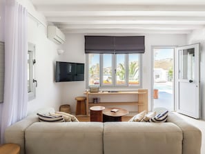Una sala de estar moderna con un sofá, una mesita de madera, un televisor montado en la pared y una ventana con vista a una piscina y edificios.