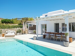 A modern outdoor pool area with a white building, a covered seating area, and a dining table with chairs. - MyMykonos Villa (Mykonos)