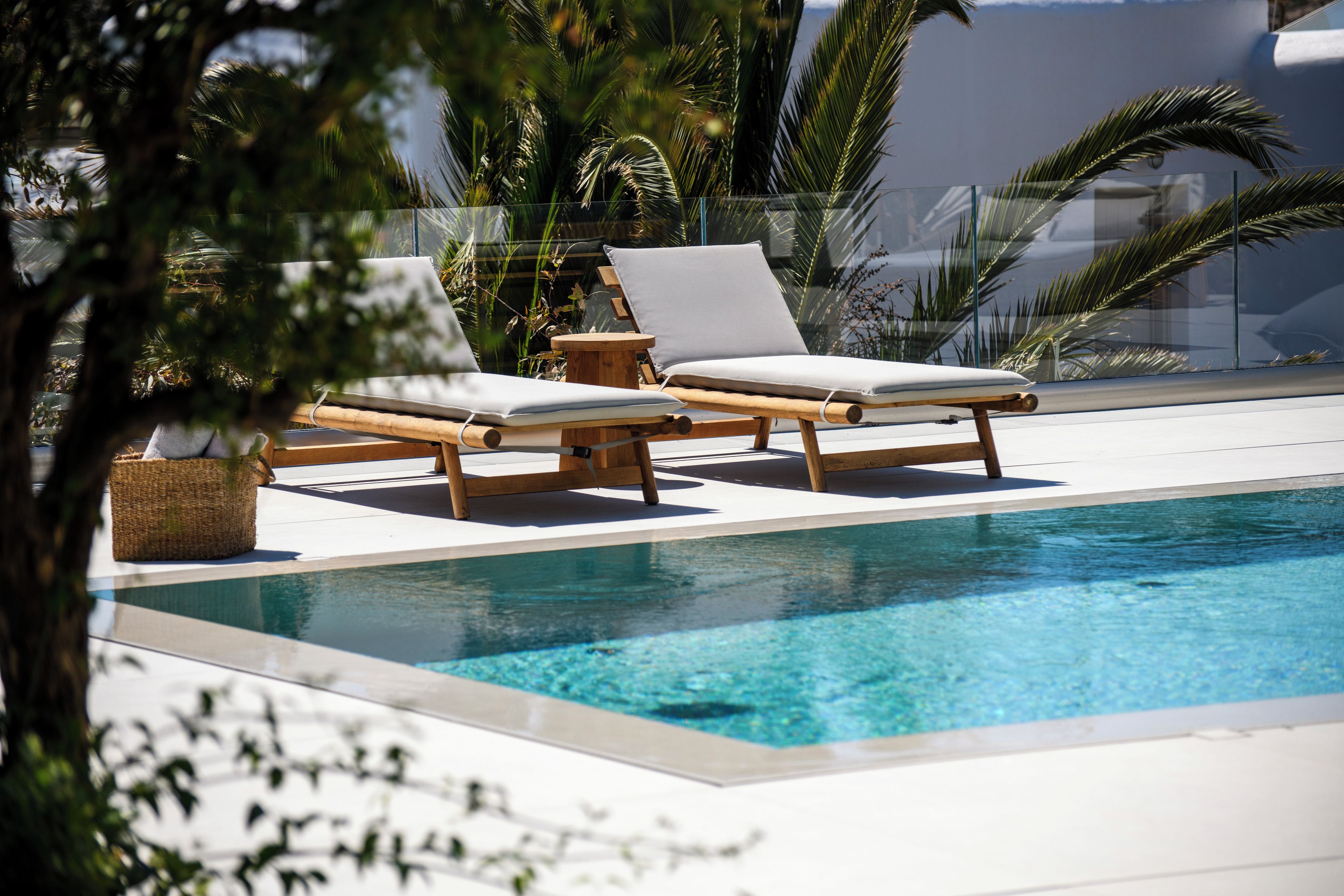 Área da piscina com duas espreguiçadeiras e uma pequena mesa, cercada por palmeiras e uma piscina azul límpida.