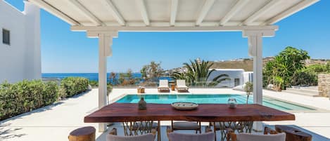 A covered outdoor seating area with a pool, white walls, and a view of the sea.