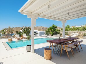 A poolside area with a table and chairs, a wooden stool, and a potted plant. - MyMykonos Villa (Mykonos)