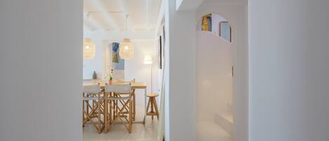 A modern interior with a dining area featuring a wooden table and chairs, pendant lights, and a white wall with a door leading to another room.