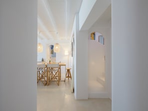 A modern interior with a dining area featuring a wooden table and chairs, pendant lights, and a white wall with a door leading to another room. - myMykonos Villa 1 (Mykonos)