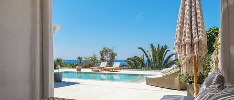 A poolside area with a view of the sea, lounge chairs, and a table.