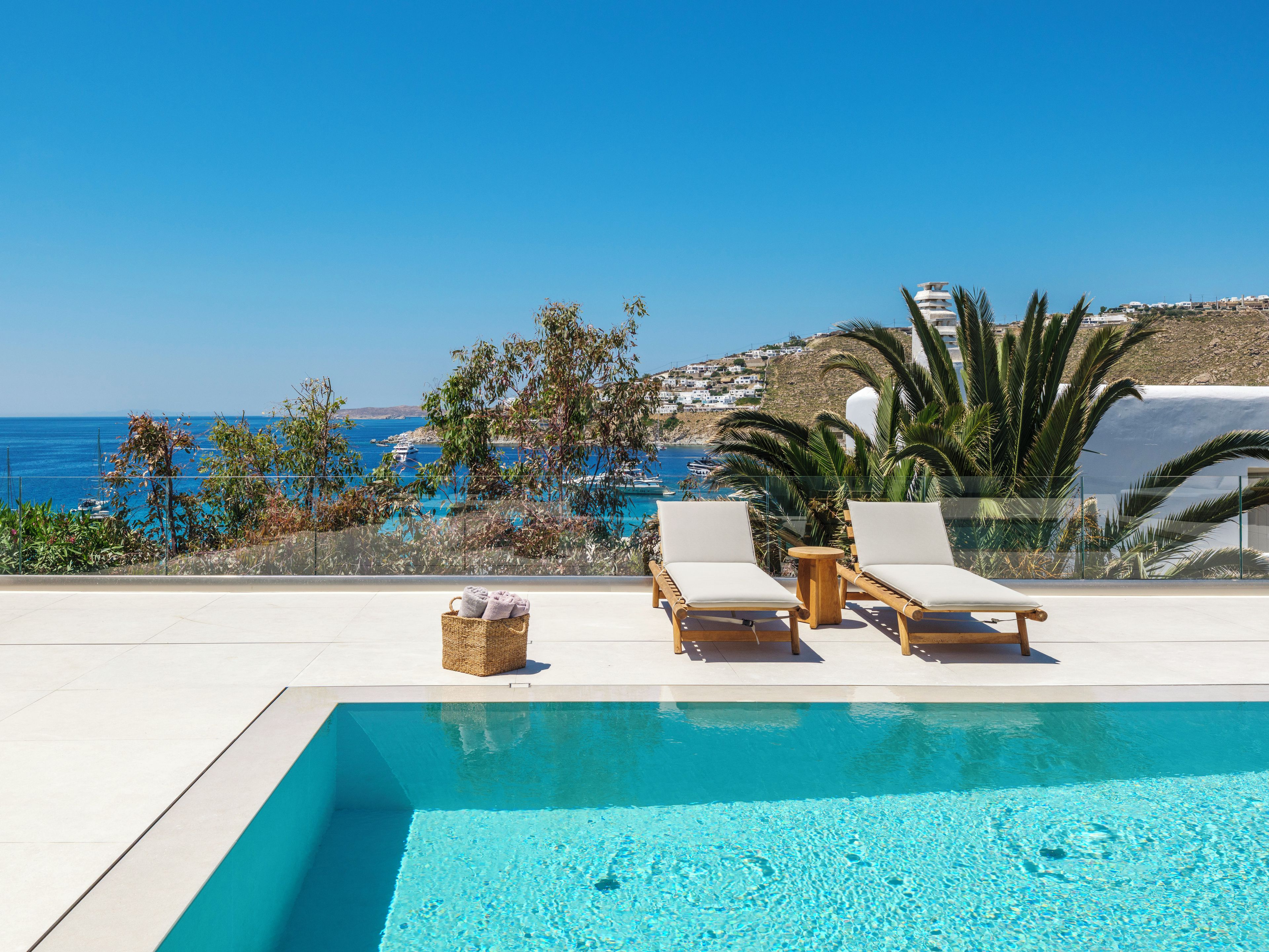 A poolside area with lounge chairs and a wicker basket, overlooking a coastal landscape with buildings and boats.