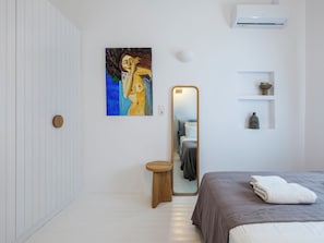 A bedroom with a bed, a bedside table, a mirror, a painting, and a shelf with decorative items. - myMykonos Villa 1 (Mykonos)