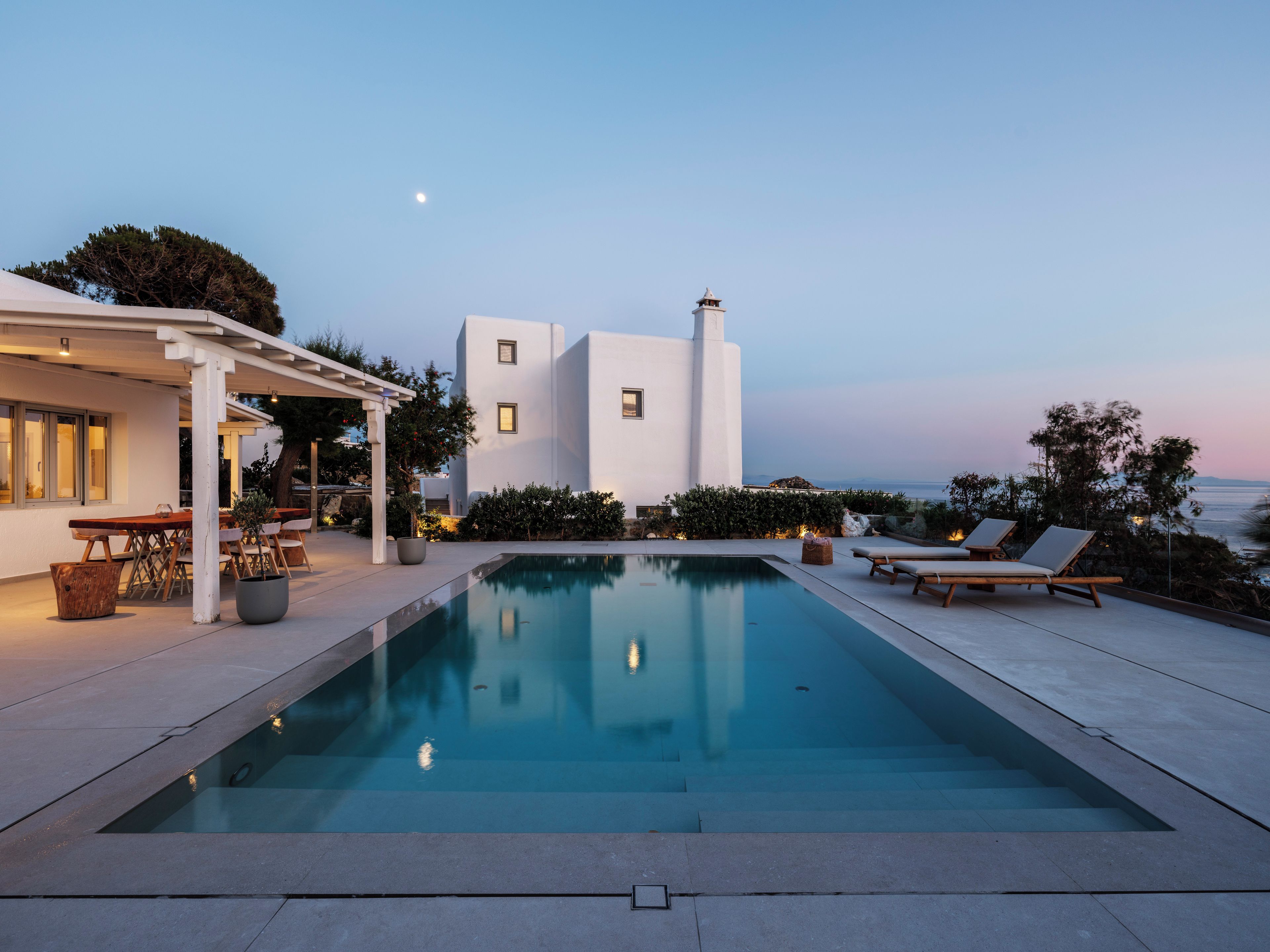 Un espace piscine moderne offrant une vue dégagée sur la mer, un bâtiment blanc et des meubles d’extérieur.