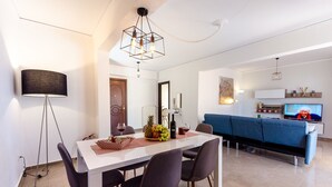 A modern living room with a white sofa, a dining table with chairs, a lamp, and a television. - Taurus - Deluxe Apartment with Sea View (Glyfada)