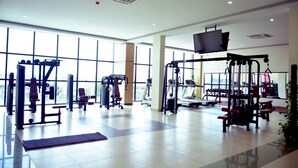 Une salle de sport spacieuse,  équipée de divers appareils d’exercice, dotée de grandes fenêtres et d’un téléviseur à écran plat fixé au mur.