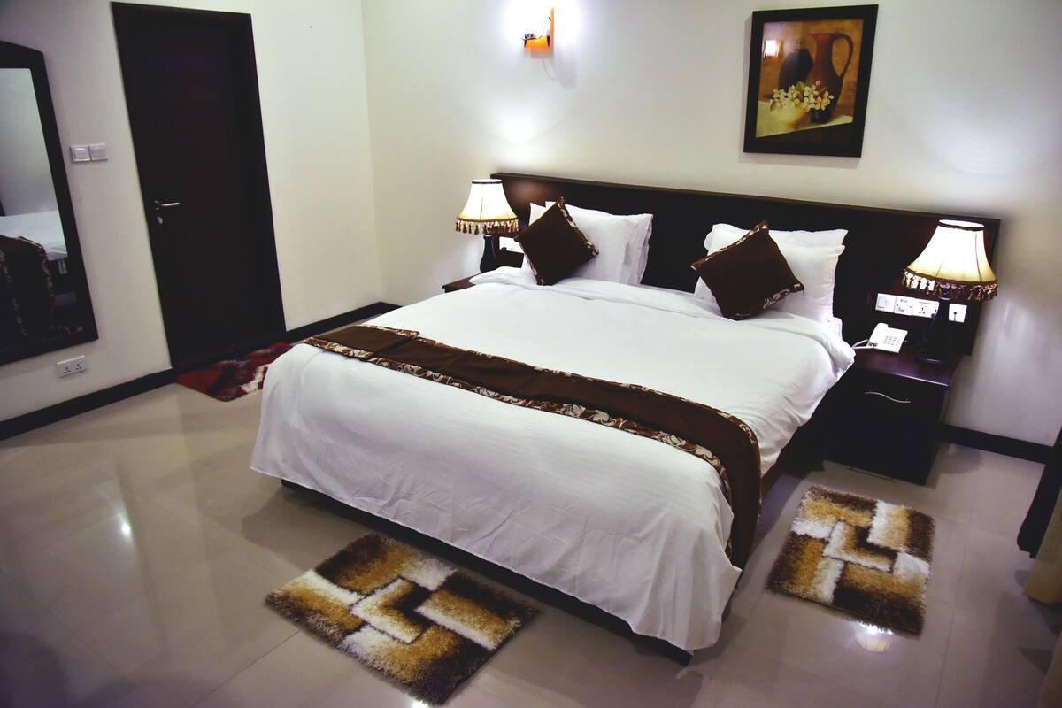 A neatly made bed with white bedding and brown accents, a nightstand with a lamp, and a wall-mounted painting.