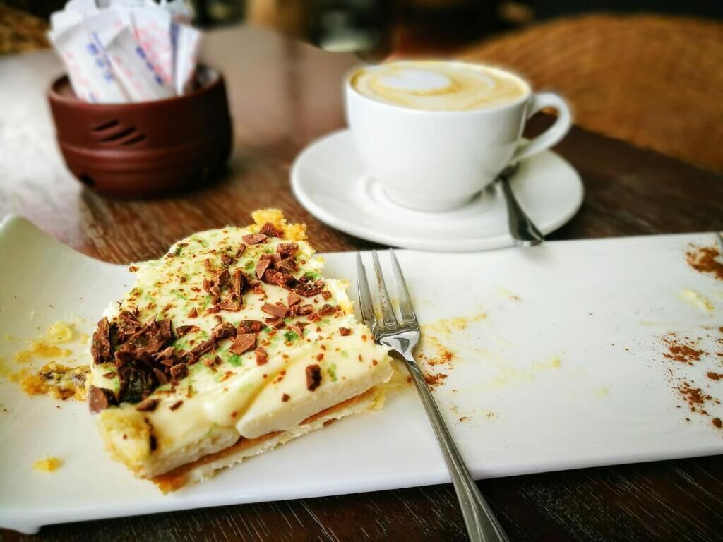 Uma fatia de cheesecake com raspas de chocolate sobre um prato branco, uma xícara de café e um garfo em uma mesa de madeira.