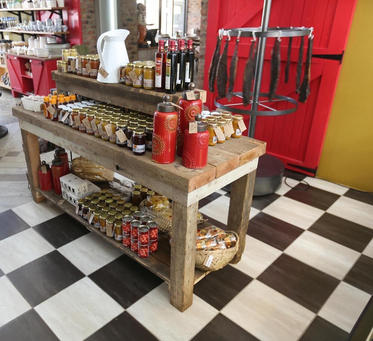 Une table d’exposition en bois, sur laquelle sont disposés divers pots et bouteilles, contenant probablement des condiments ou des conserves, dans un magasin dont le sol est à carreaux et les murs sont rouges.